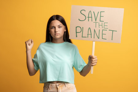 Serious confident brunette teen girl showing holding save the planet poster on yellow background.の写真素材