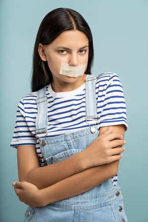 Sad teenage girl with mouth sealed with tape. Restriction of freedom of speech, authoritarianismの写真素材