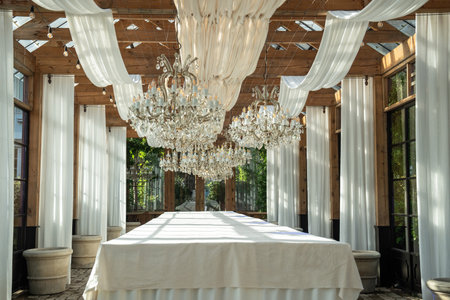 Long table covered with white tablecloth stands under canopy and luxury chandeliers on sunny dayの写真素材