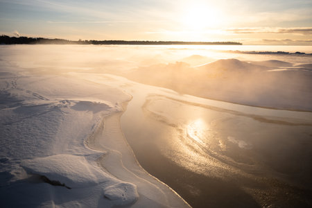 Icy lake with sunlight reflecting off smooth surface. Mist rising from unfrozen snow covered water
