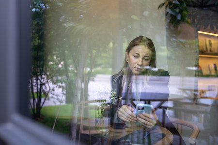 View through glass relaxed woman scrolling news or social media on phone taking coffee break in cafe