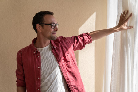 Smiling man holding on to curtain and looking out window in summer weather admiring cityscape