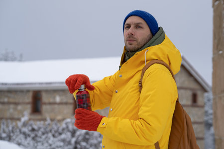 Thoughfull man in a yellow jacket holds thermos with hot drink in hands on snow winter day outdoors.