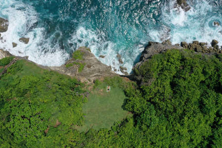Aerial photo of man lying on a green cliff above the blue Indian Ocean and foaming breaking waves,...