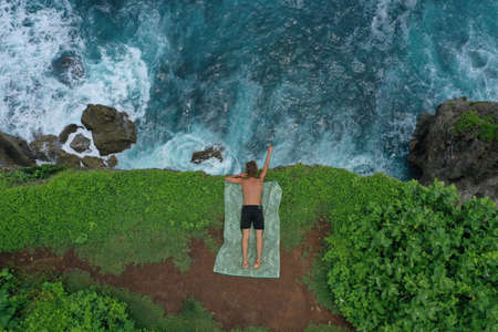 Aerial photo of man lying on a green cliff above the blue Indian Ocean and foaming breaking waves,...