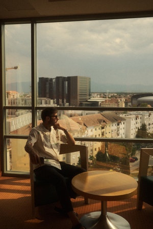 A man sits peacefully by a tall window, gazing at a cityscapeの写真素材