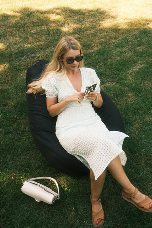 A woman in a white dress sits on a bean bag, looking at her phoneの写真素材