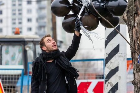 2015.11.22, Moscow, Russia. birthday is a sad holiday concept. A young man wearing black scarf and coat holding black balloons on background of urban scene, blur and grain effect.の写真素材