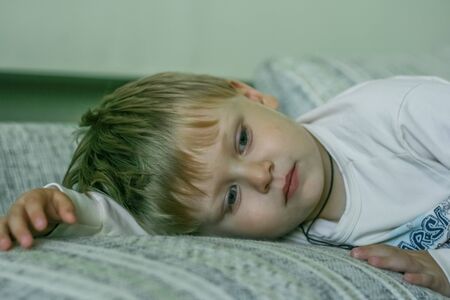 portrait of a little fair-haired sad boy lying on a sofa and watching TV.