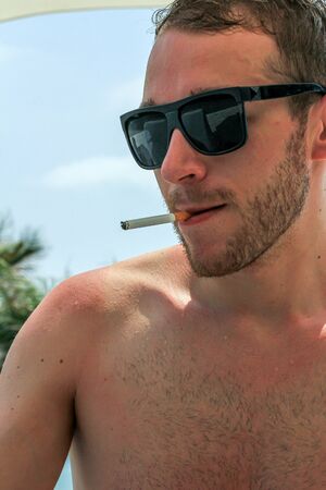 young guy with wet hair and black glasses smiking a cigarette on the beachの写真素材