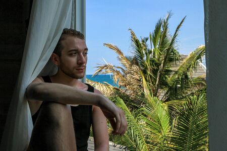 young bearded guy is resting sitting on the windowsill and looking at the sea