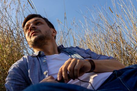 a young attractive man wearing jeans, white T-shirt and a blue shirt lies on a field of cereals against a blue sky and looks away to the leftの写真素材