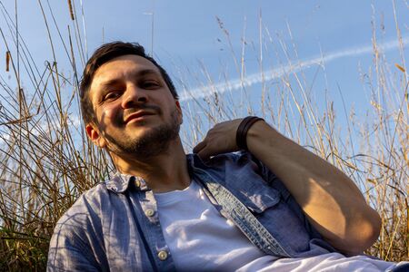 a young attractive man wearing jeans, white T-shirt and a blue shirt lies on a field of cereals against a blue sky and looks to cameraの写真素材