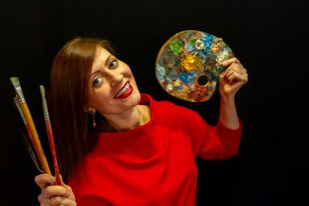 A young beautiful woman wearing red dress on black background. pretty young smiling woman holding a palette and artistic brushes.の写真素材