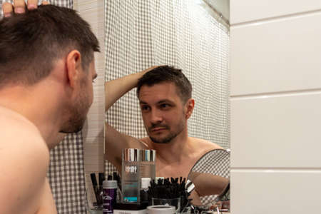 05/19/2018, Moscow, Russia. young short-haired guy with unshaven face looks in the mirror in the bathroom.のeditorial素材