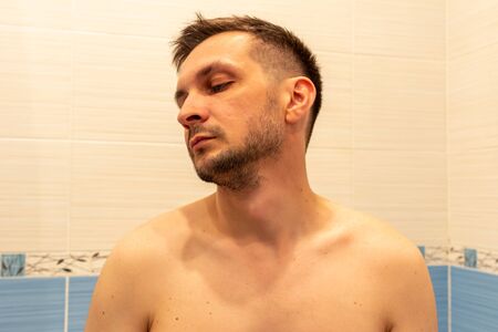 young attractive shirtless man sitting in the bathroom on the background of a light tile and looking away.の写真素材