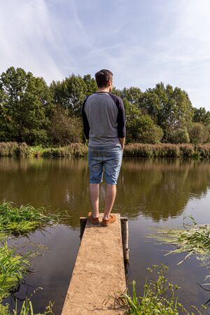 A young guy in denim shorts and a longsleeve stands on a wooden structure by the river, with his hands in his pockets and looking at the river, back and side view.の写真素材