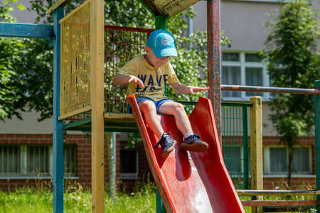 06/30/2020, Maloyaroslavets, Russia. little fair-haired boy wearing a blue baseball cap rolls down a children's slide on a playground. A kid having fun in the yard.のeditorial素材