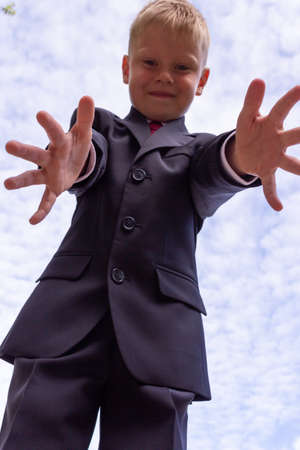Portrait of a blond first-grader boy dressed in a dark suit with his arms outstretched to the camera on background of cloudy sky. A boy having fun, blur and grain effect.の写真素材