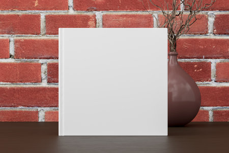 Square book cover mock up standing on a dark wooden desk with red brick wall background. 3d illustrationの写真素材