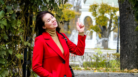 Close up outdoor portrait of Young pretty woman at the autumn park. girl wearing red coat points to the up. Fashion Art design with copy space for your text. Cute lady, golden autumn background.の写真素材
