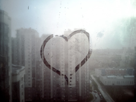 symbol of the heart and love is drawn on a wet misted glass in the sun. selective focus. Heart on fogged glass and hand. Romantic background for valentines day. Big City buildings on the backgroundの写真素材