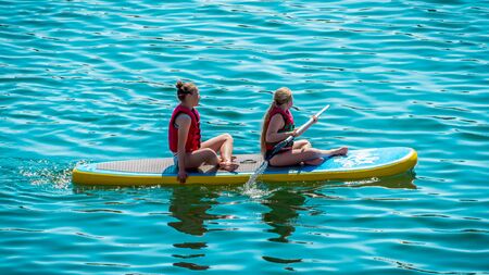 TALLINN, ESTONIA- JUNE 02 , 2018:  Wakepark on the Manniku lake. view of 2 young girls exercising with sup board in turquoise tropical. Sittiing and paddling on  water. SUP (standup paddleboarding)のeditorial素材