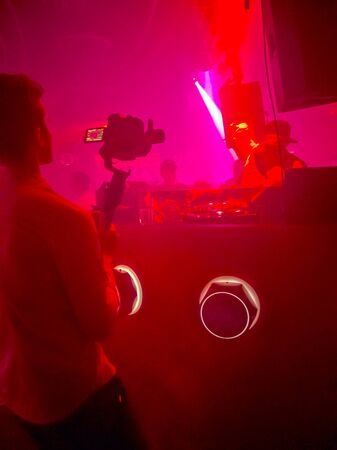 Silhouette of a camera operator taking video or photo and DJ from back playing in nightclub in front...