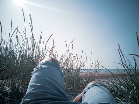 First-person view of a man relaxing, enjoying view over the beach with wild grasses. legs in jeans of hidden man lying behind a grass dune. Grass growing on the hill and sea surface. Vacation conceptの写真素材