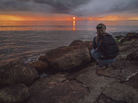 Happy young joyful woman traveler photographer with a backpack and camera during taking photos of sea at sunset. Baltic sea, Tallinn, Estoniaの写真素材