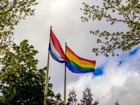LGBT and Holland flags on poles blow in the wind. Symbol of tolerant. Gay sign rainbow. The rainbow pride, Trees and blue cloudy sky on the background. Copy spaceの写真素材