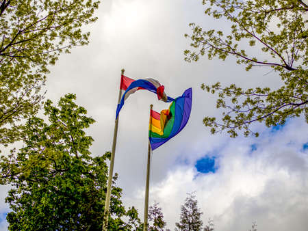 LGBT and Holland flags on poles blow in the wind. Symbol of tolerant. Gay sign rainbow. The rainbow pride, Trees and blue cloudy sky on the background. Copy spaceの写真素材