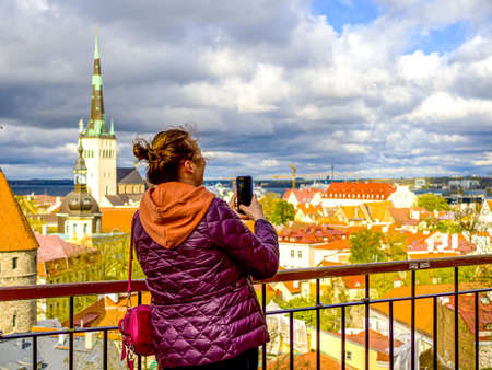 Tourist in Estonia, taking photo of old town with smartphone. Travel photography with mobile phone. travel and photography concept, young beautiful woman in red looks towards city towers. copy spaceの写真素材