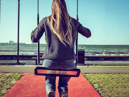 Back view of young girl on swing at ocean beach during sunny day in Estonia, Tallinn. Silhouette of happy young woman on a swing with sea and blue sky background. Toned image. Summer and vacation concept. Copy spaceの写真素材
