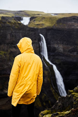 A young guy stands on a cliff and looks at the waterfall. The guy in the yellow raincoat.の写真素材