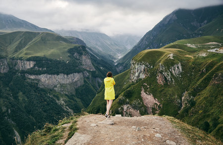 A young Girl in a yellow raincoat tourist photographs the mountains. Girl making a photo shoot of...