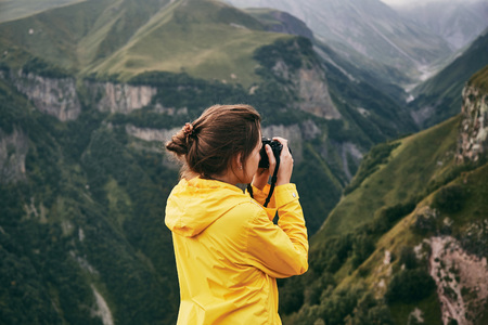 A young Girl in a yellow raincoat tourist photographs the mountains. Girl making a photo shoot of...