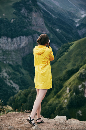 A young Girl in a yellow raincoat tourist photographs the mountains. Girl making a photo shoot of...