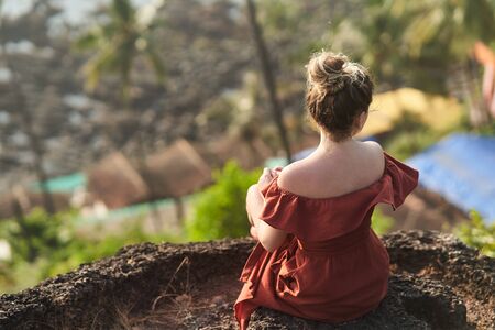 A young girl in a red dress sits on a rock and looks at the landscape.の写真素材