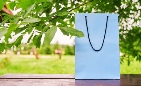 Empty paper bag stands on a wooden table against the background of green leaves. Shopping concept. High-quality photo.の写真素材