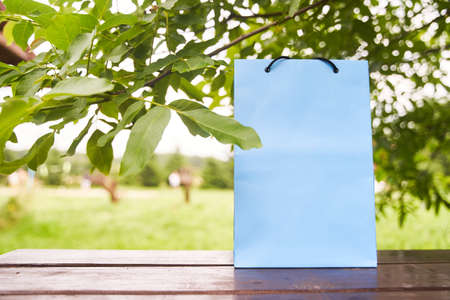 Blank paper bag stands on a wooden table against the background of green leaves. Shopping concept. Copy space.の写真素材