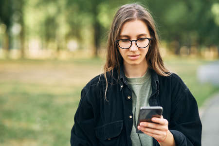 Young woman with glasses reading a message on her phone while standing in the park