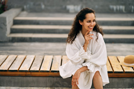 Smiling curly brunette girl in a white dress sitting on a wooden bench in the parkの写真素材