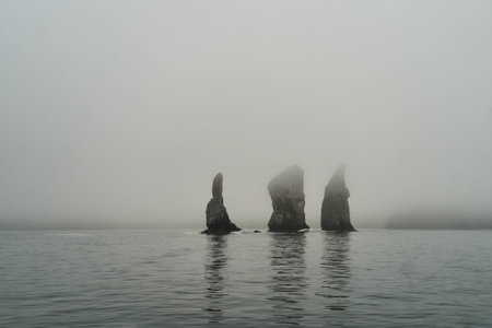 Mysterious and Fog-Shrouded Rock Formations Rising Over Calm and Serene Water Surfaceの写真素材