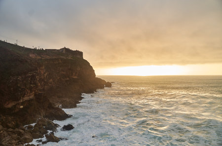 View of the sunset and the old lighthouse on a rock with a fortress on the coast of the Atlantic Ocean in the city of Nazare, Portugal.の写真素材
