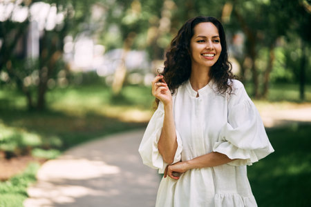 Portrait of a beautiful curly-haired brunette girl in the park. Girl smiling and holding her hairの写真素材