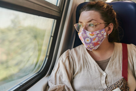 A girl in glasses and a medical mask riding a train and looking out the window