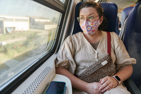 A girl in glasses and a medical mask riding a train and looking out the window