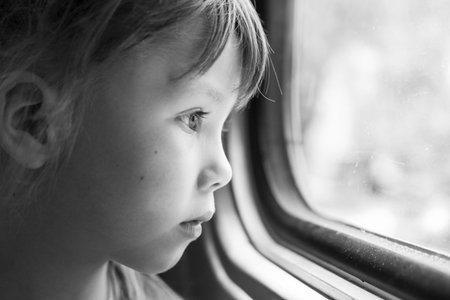 Monochrome portrait of a beautiful girl who looks in the window of the train. Close-up of a sad child looking through window. Black and white photographyの写真素材