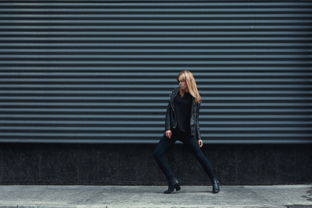 Young beautiful pretty blonde european girl walking along the street wearing black leather jacket, black jeans. Dark metal background. Trendy, Urban street style. Autumn Vogue Style. Toned ifiltersの写真素材